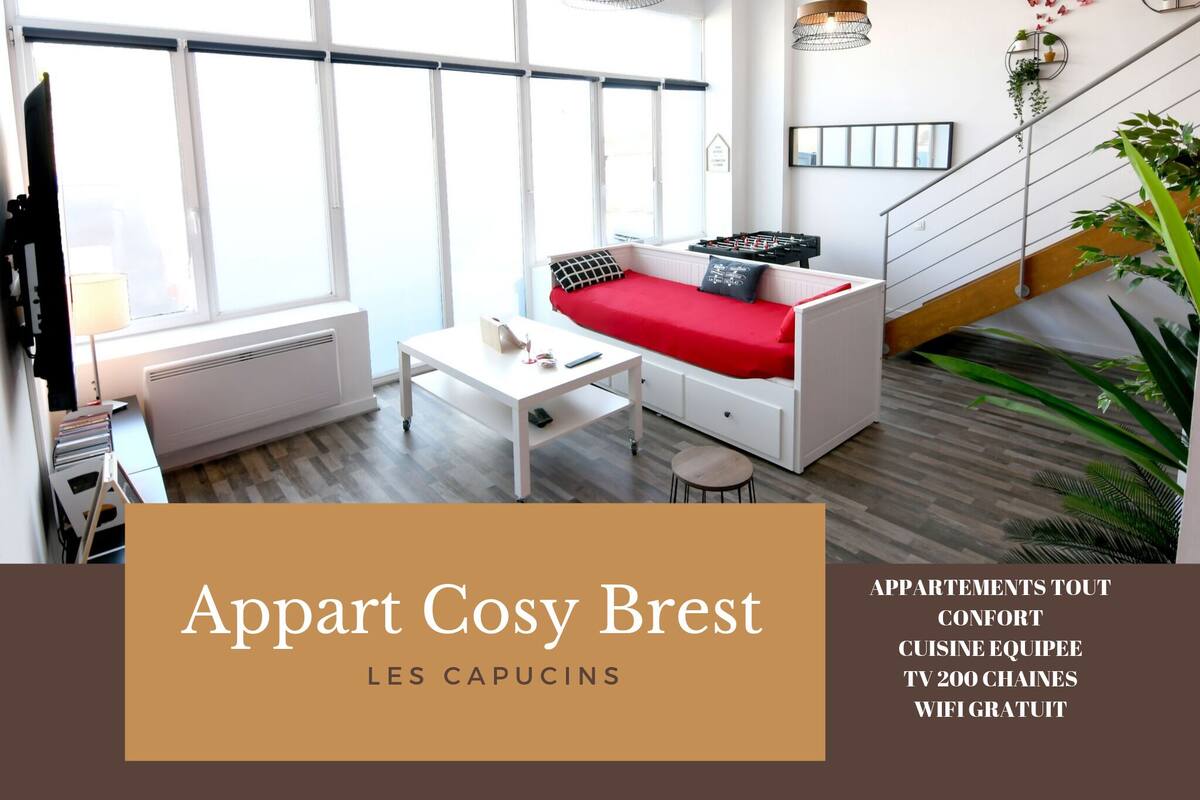 A modern apartment with a sofa, a small table, and a TV. It has large windows, a staircase, and potted plants.