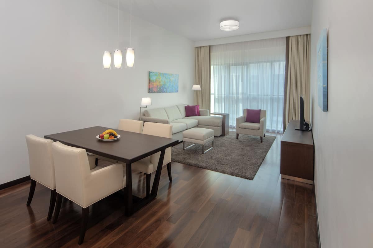 A modern hotel room with a dining area, a sofa set, a flat-screen TV, and a large window with curtains.