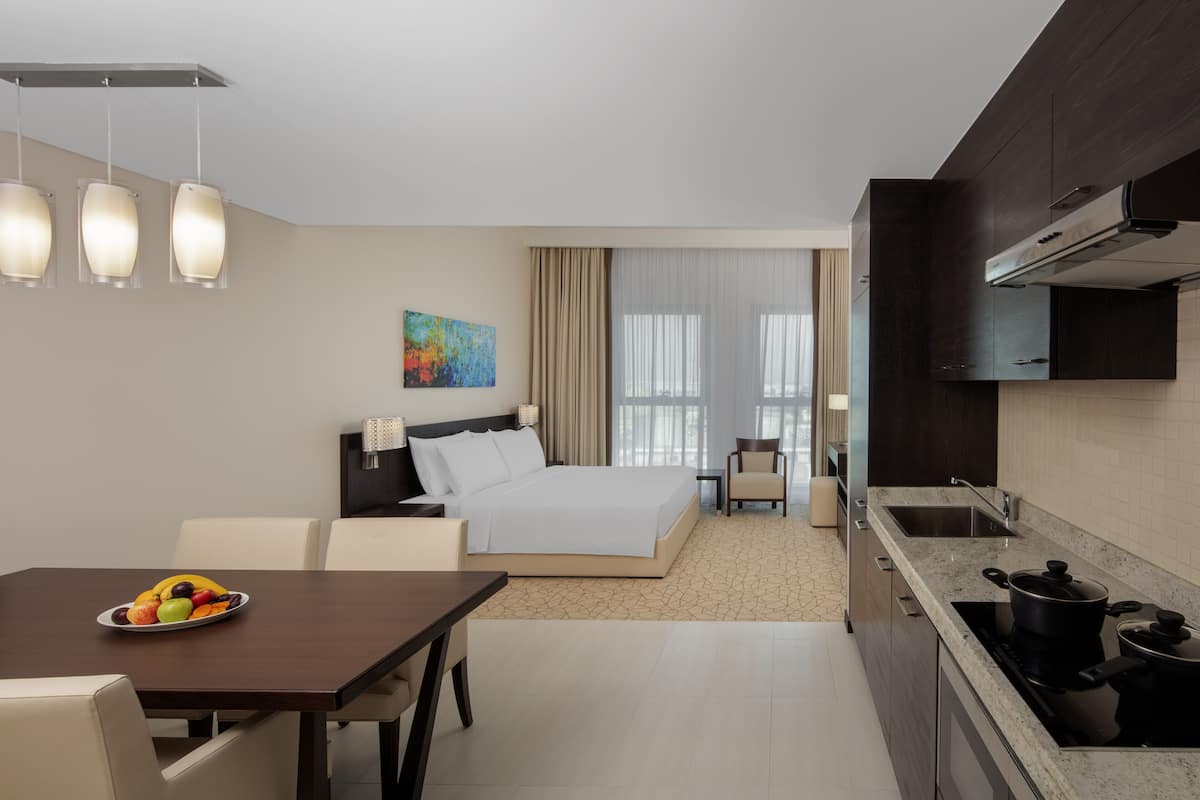 A hotel room with a bed, bedside table, and a dining area with a fruit bowl.