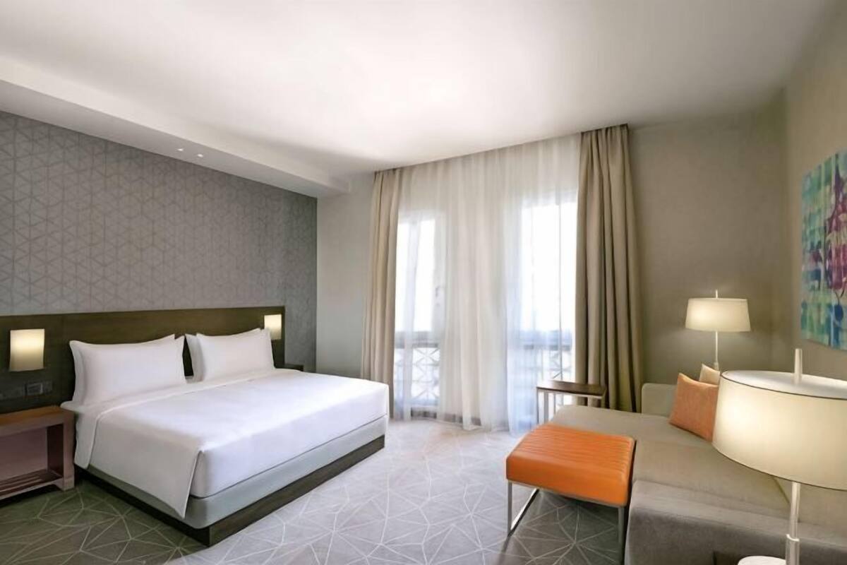A hotel room with a bed, a desk, a chair, a lamp, and a view of the outdoors.