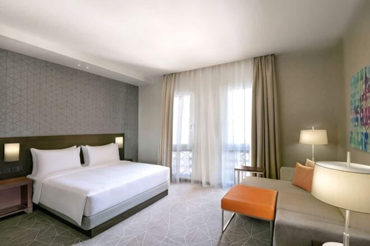 A hotel room with a bed, a desk, a chair, a lamp, and a view of the outdoors.