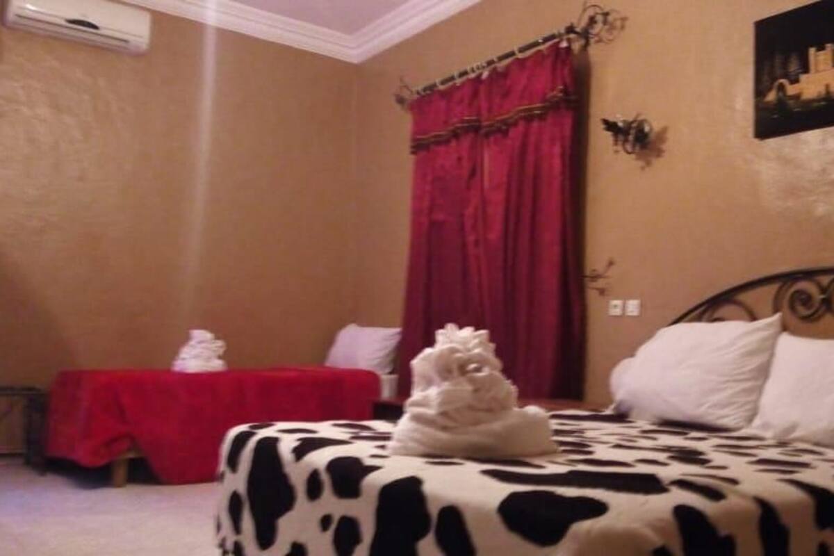 A bedroom with a bed covered in a cow print blanket, a red table with a white towel, and a red curtain.