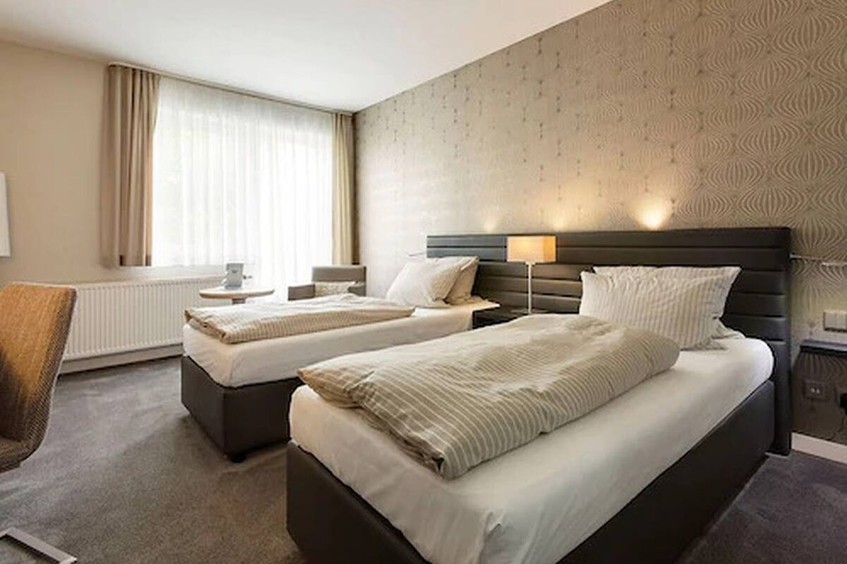 Quarto de hotel com duas camas, uma cadeira, uma mesa e uma janela com cortinas.