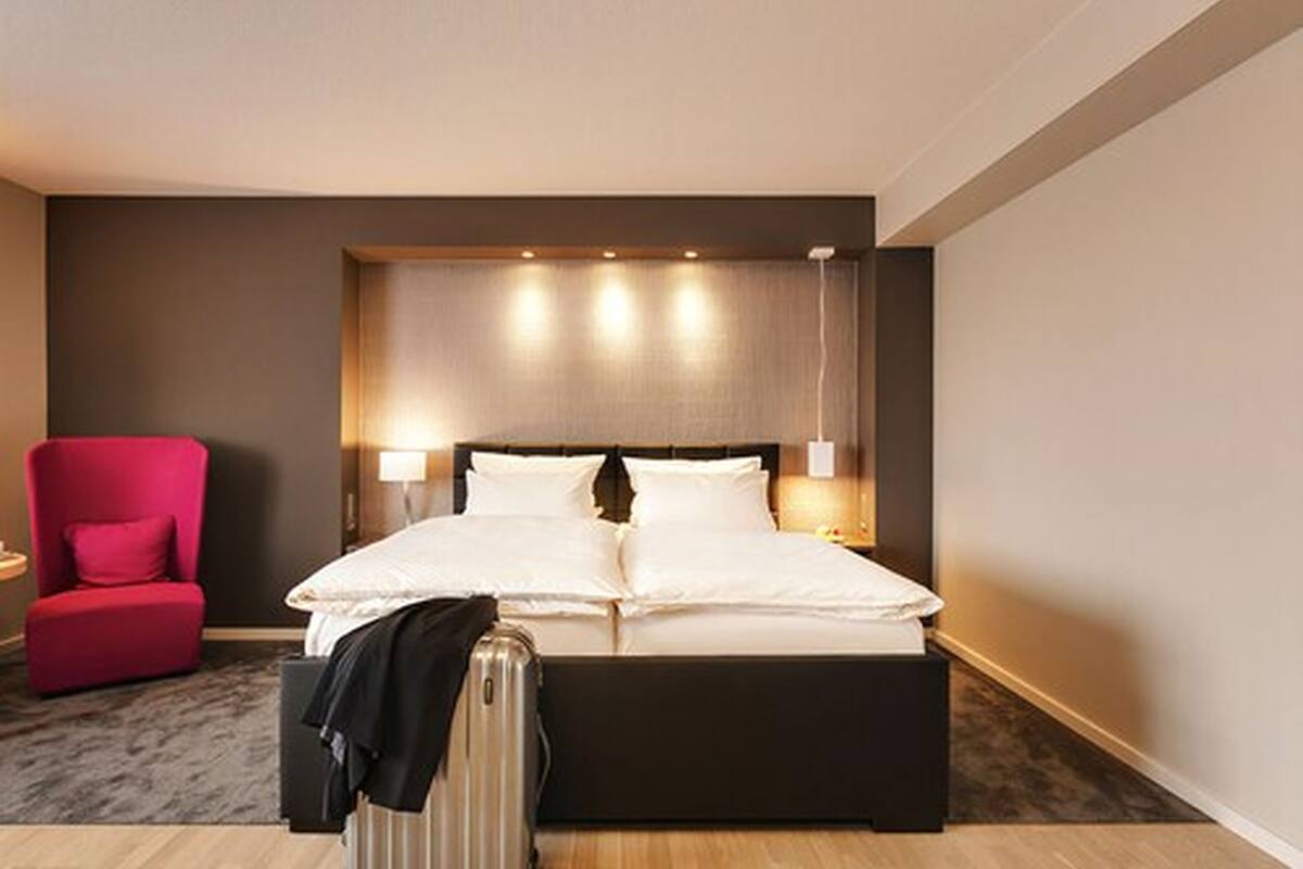 Quarto de hotel moderno com uma cama grande, uma poltrona vermelha e uma mala no chão.