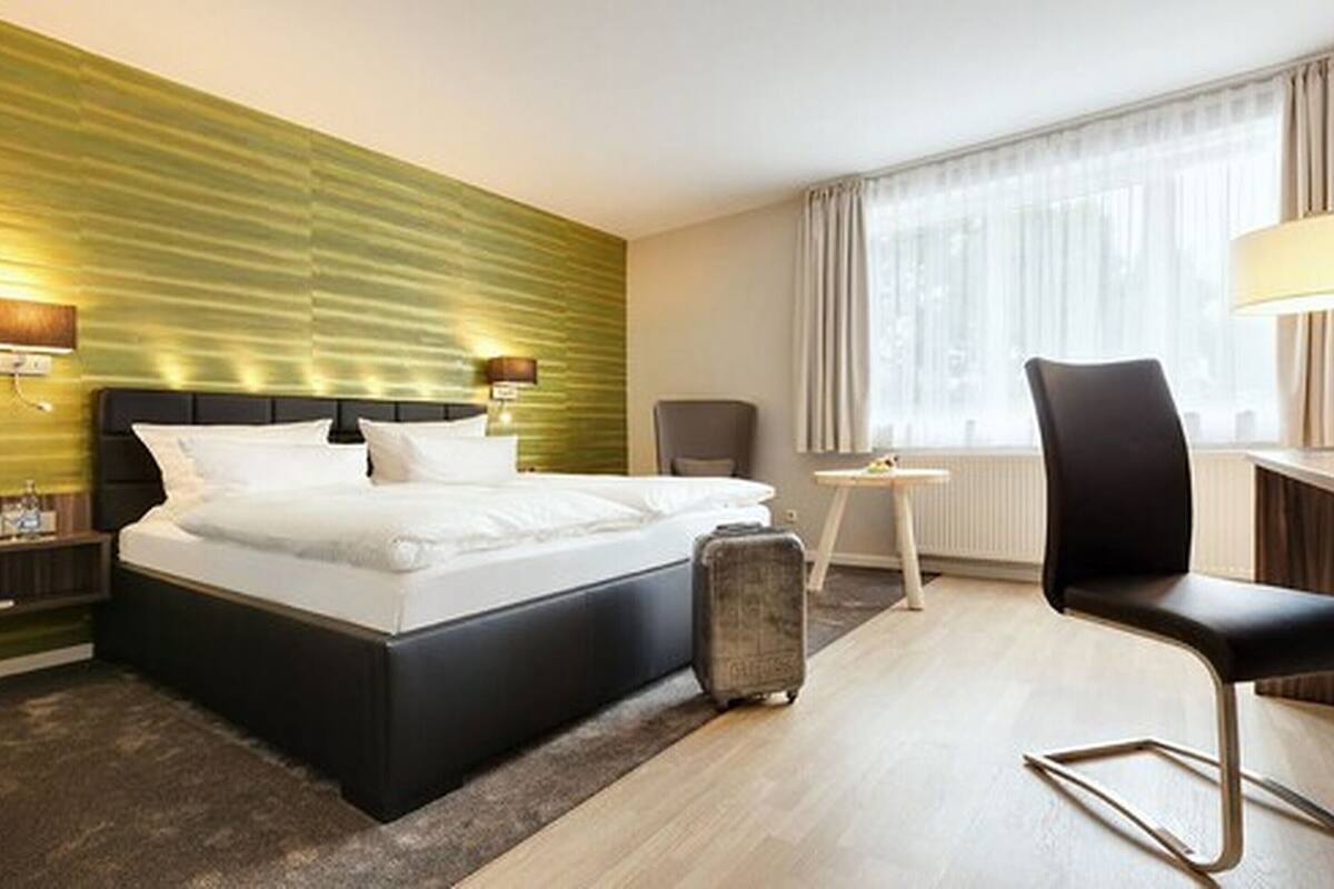 Quarto de hotel moderno com uma cama grande, uma mala, uma escrivaninha com cadeira e uma janela com cortinas.