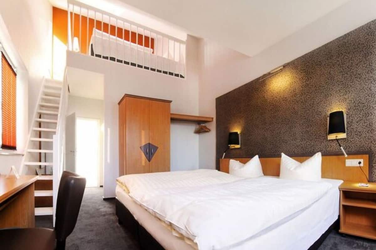 Quarto de hotel com cama, secretária, cadeira, guarda-roupa, escada e luminária fixada na parede.