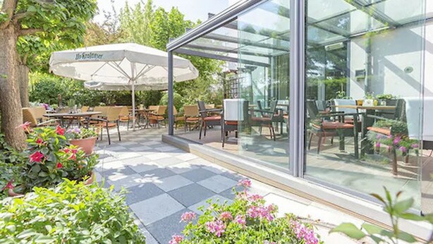 Een terras buiten met tafels en stoelen, een glazen overkapping en potplanten.