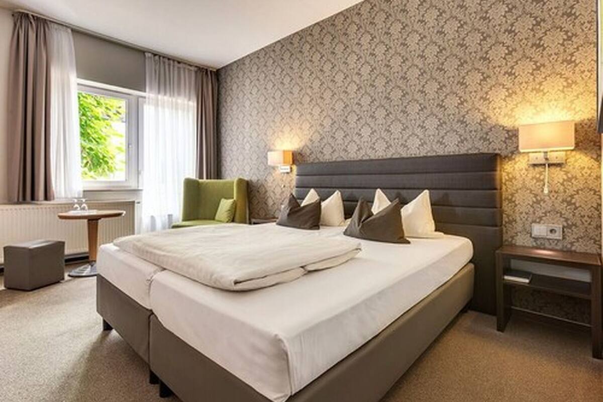Quarto de hotel com uma cama grande, uma mesa pequena, uma cadeira e uma janela com cortinas.