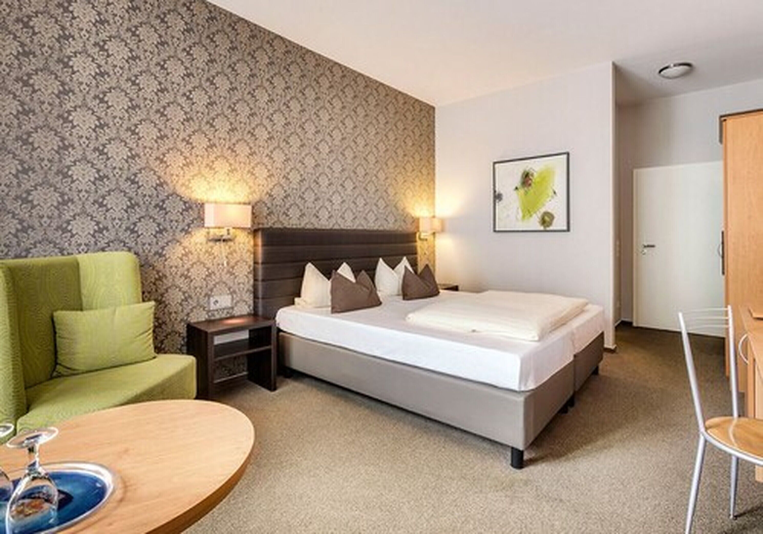 A hotel room with a bed, a green armchair, a bedside table with a lamp, and a framed picture on the wall.
