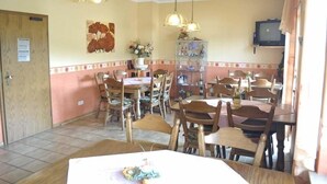 Zona pranzo con tavoli e sedie in legno, televisione a parete e lampade a sospensione.