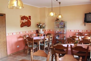 Un comedor con mesas y sillas de madera, un televisor montado en la pared y lámparas colgantes.