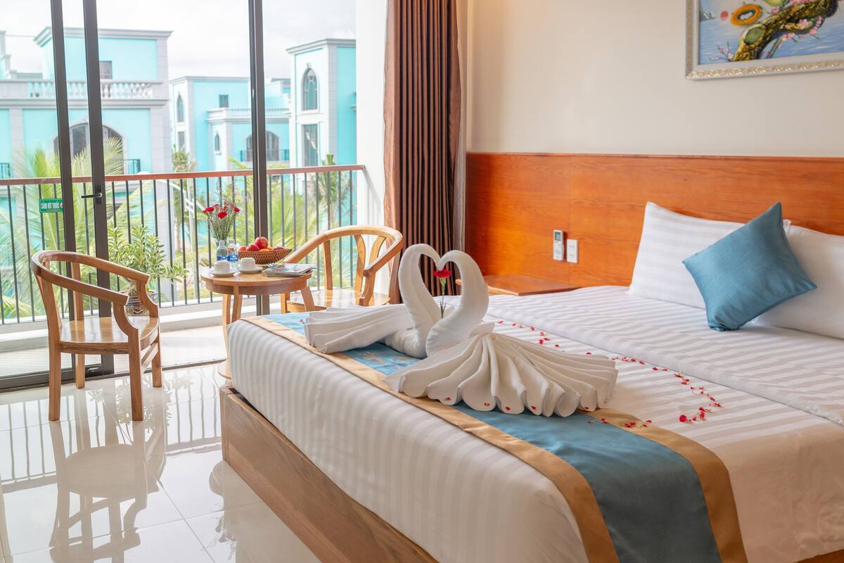A hotel room with a bed, a wooden headboard, a balcony with a view of buildings, a chair, and a table with flowers.