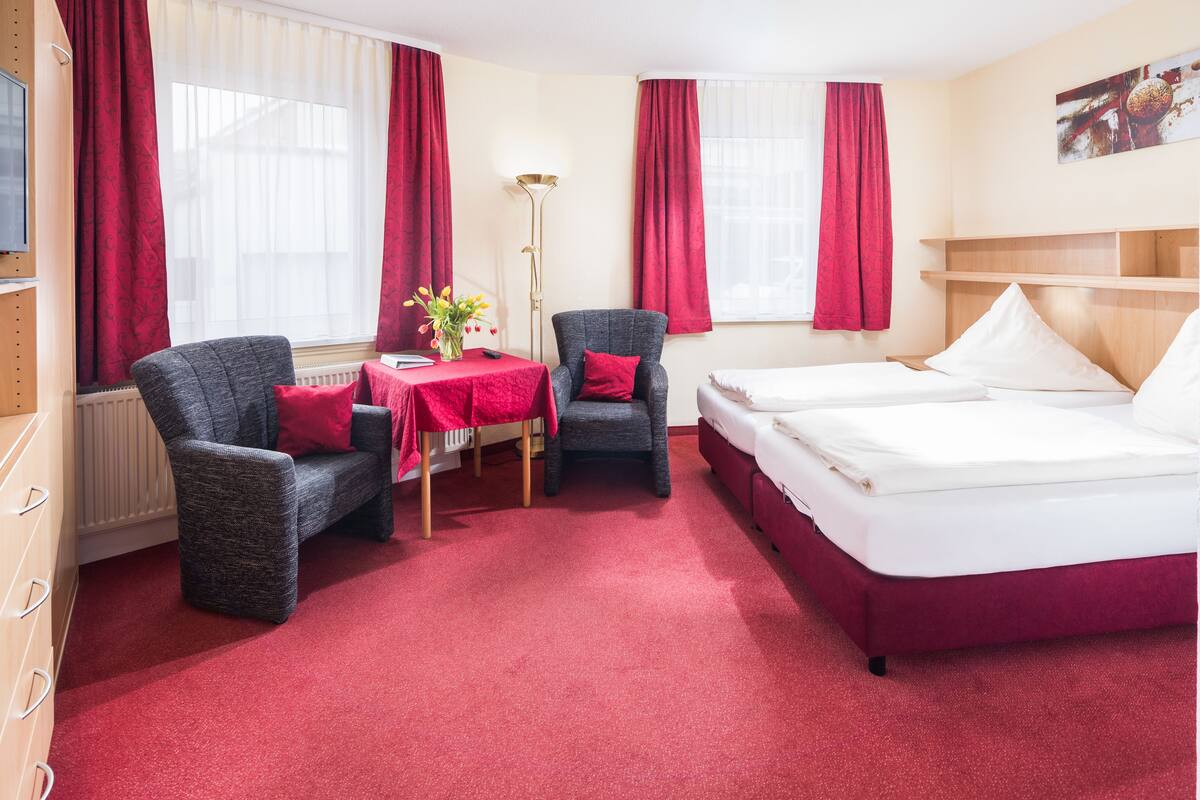 A hotel room with two single beds, a desk with a chair, a red carpet, and red curtains.