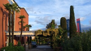 A hotel entrance with a covered walkway, palm trees, and a car parked in front. - Hapimag Resort Ascona (Ascona)