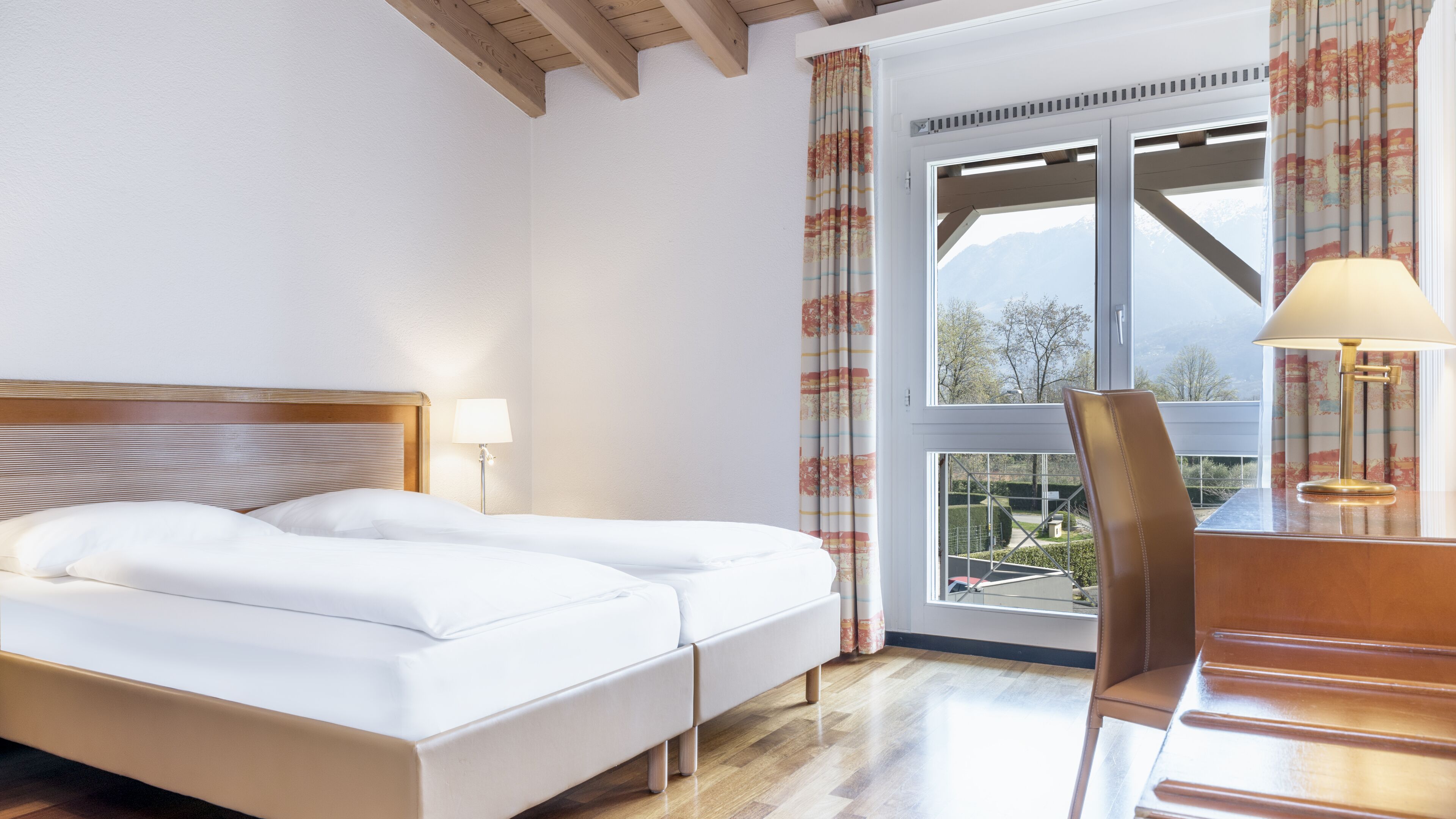 Ein Hotelzimmer mit Bett, Schreibtisch mit Lampe und Blick ins Freie durch ein Fenster.
