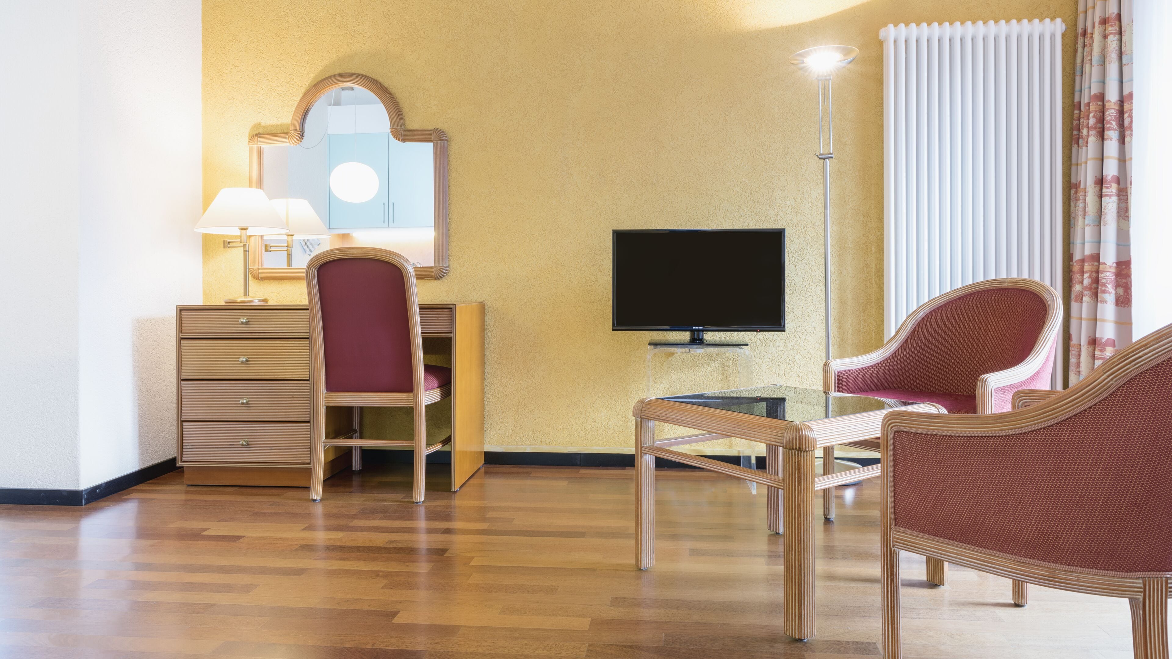 A hotel room with a wooden floor, a desk with a lamp, a chair, a television, and a large window with curtains.