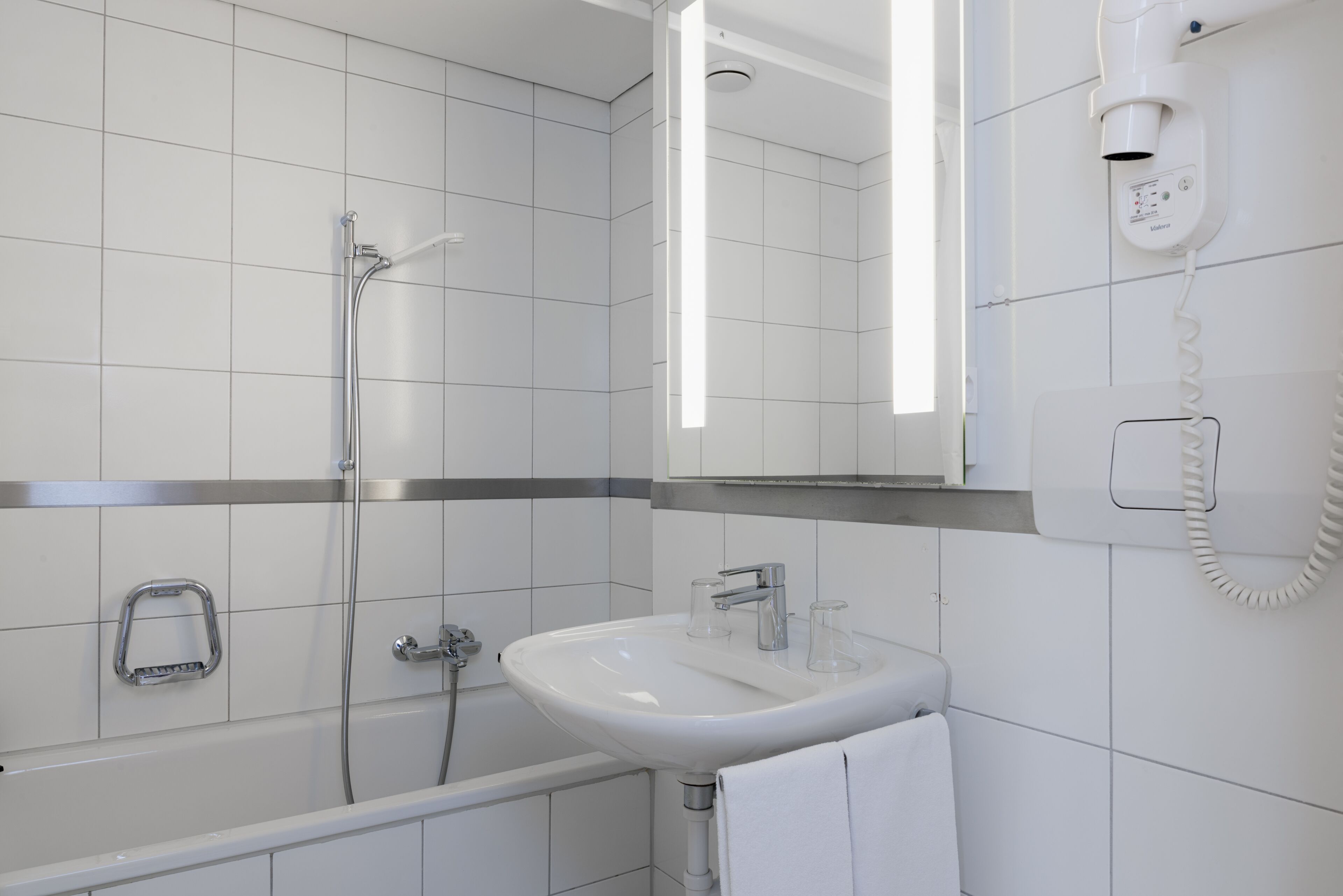 A modern bathroom with white tiles, a bathtub, a sink, a mirror, and a hairdryer mounted on the wall.