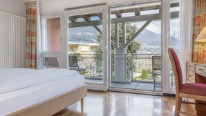 A bedroom with a bed, a chair, a table, and a balcony with a mountain view. - Hapimag Resort Ascona (Ascona)