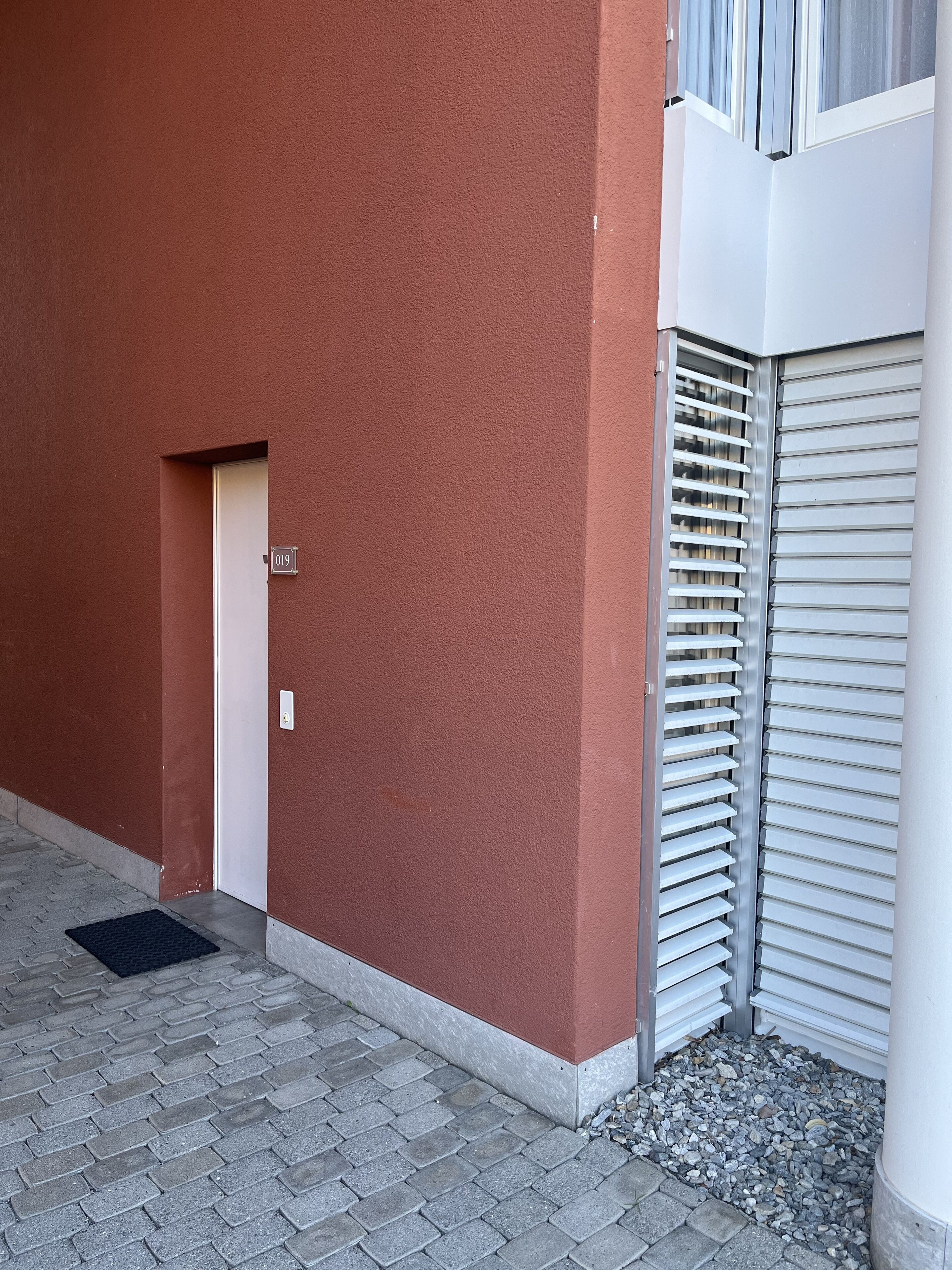 Um edifício com fachada vermelha, uma porta com placa de número e uma janela com venezianas.