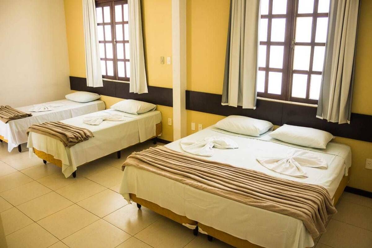 Quarto de hotel com três camas individuais, cada uma com colcha branca e manta marrom. O quarto tem uma parede de destaque amarela e janelas amplas com cortinas.