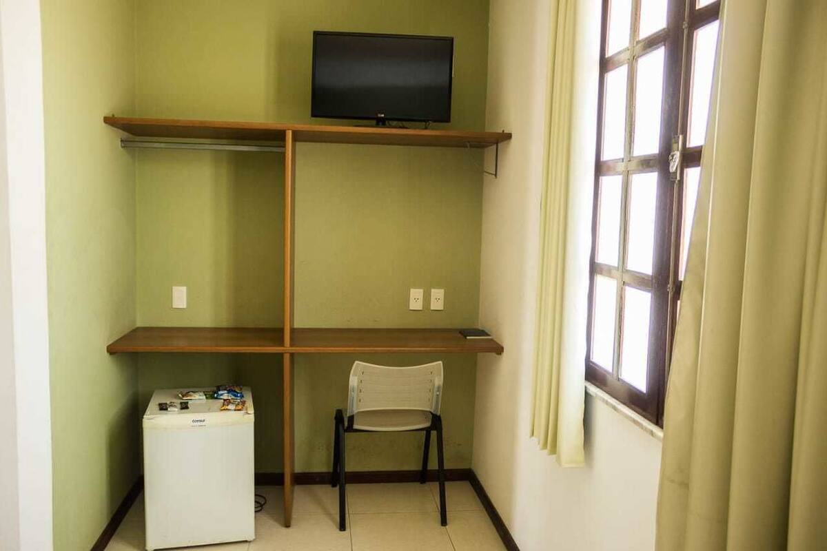 Quarto pequeno com uma cama de solteiro, mesa de cabeceira, janela com cortinas e televisão presa na parede.