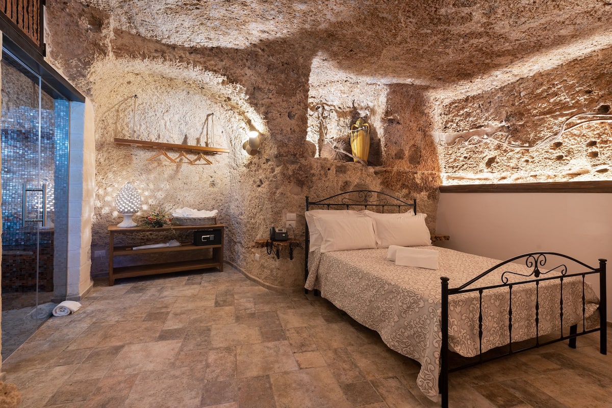 A bedroom with a stone wall, a bed, a nightstand, a lamp, and a view of the city.