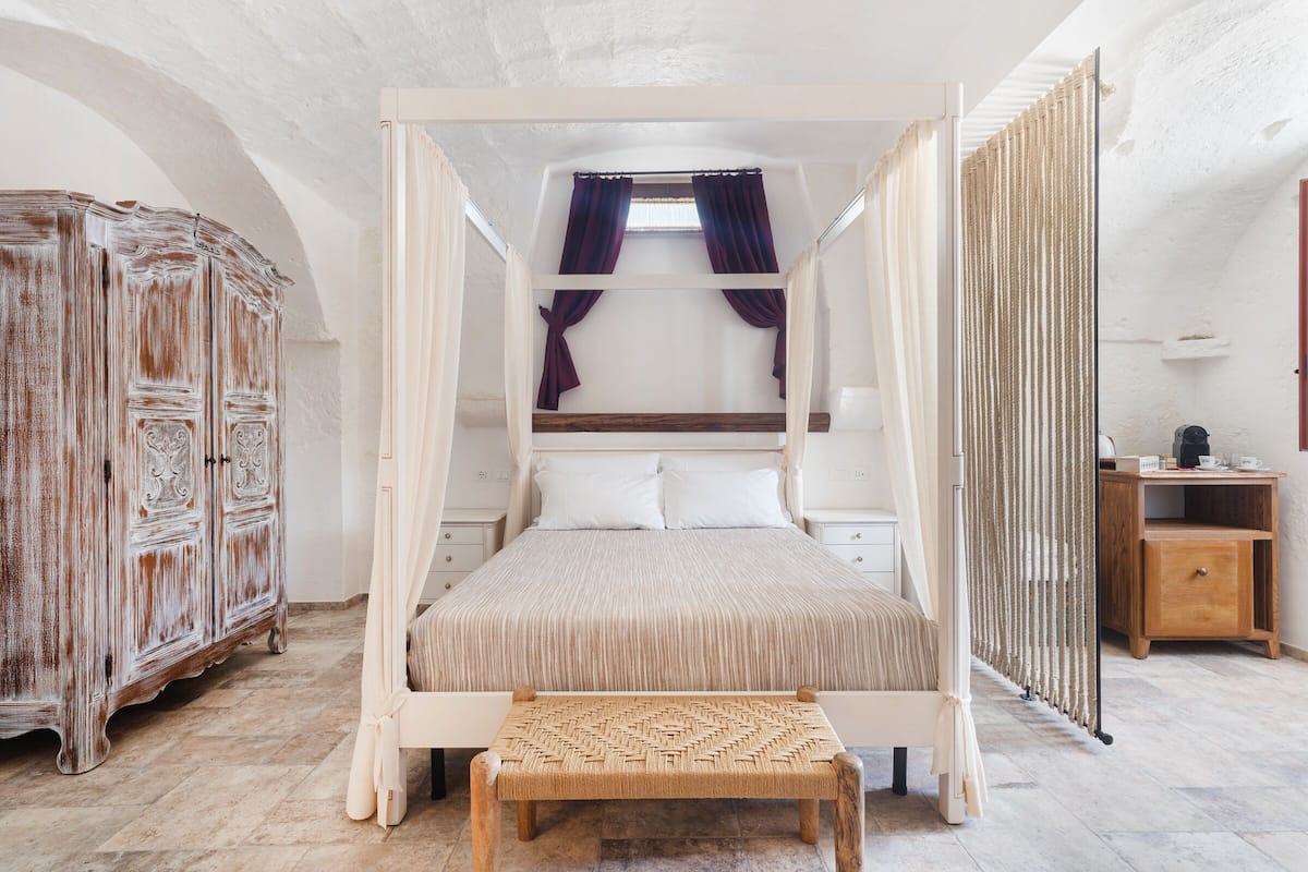 A four-poster bed with a woven bench in front, a wooden cabinet to the left, and a small wooden table to the right.