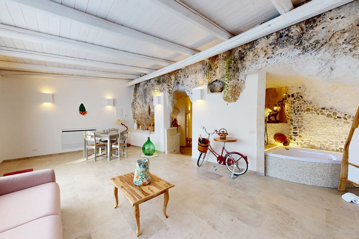A spacious living area with a dining table, a sofa, and a wooden coffee table. A red bicycle is parked against the wall, and there is a stone wall with a rustic aesthetic.