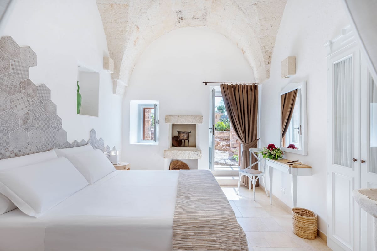A bedroom with a large bed, a small table with flowers, a view of the outside, and a rustic ceiling.