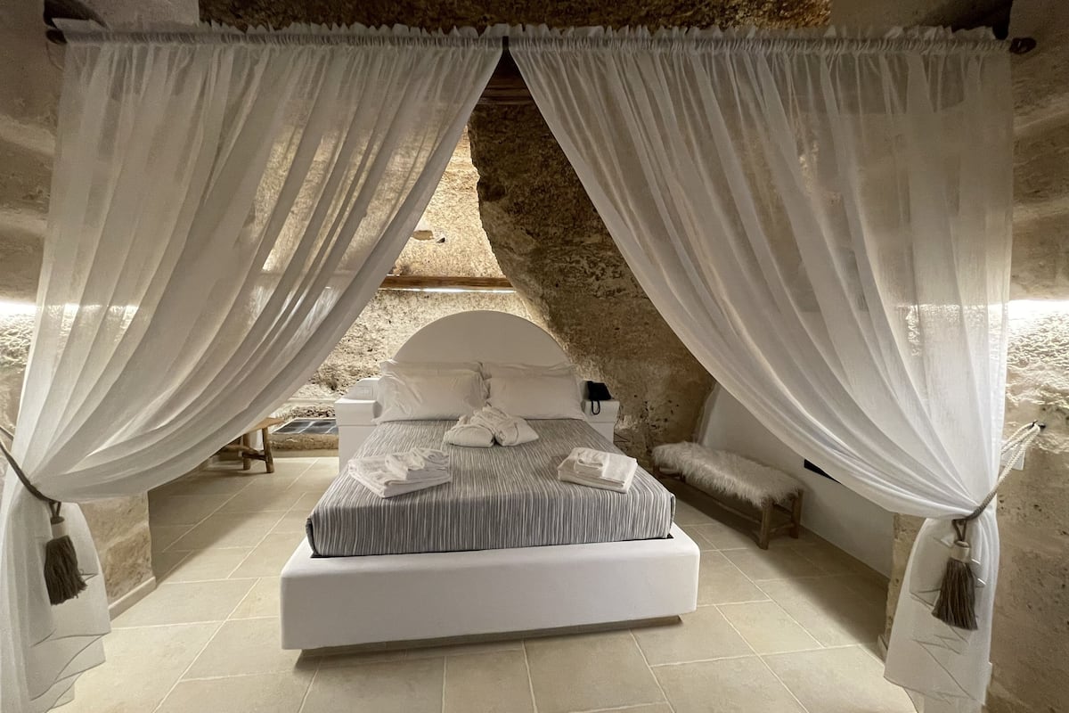 A bedroom with a bed, white curtains, and a stone wall.