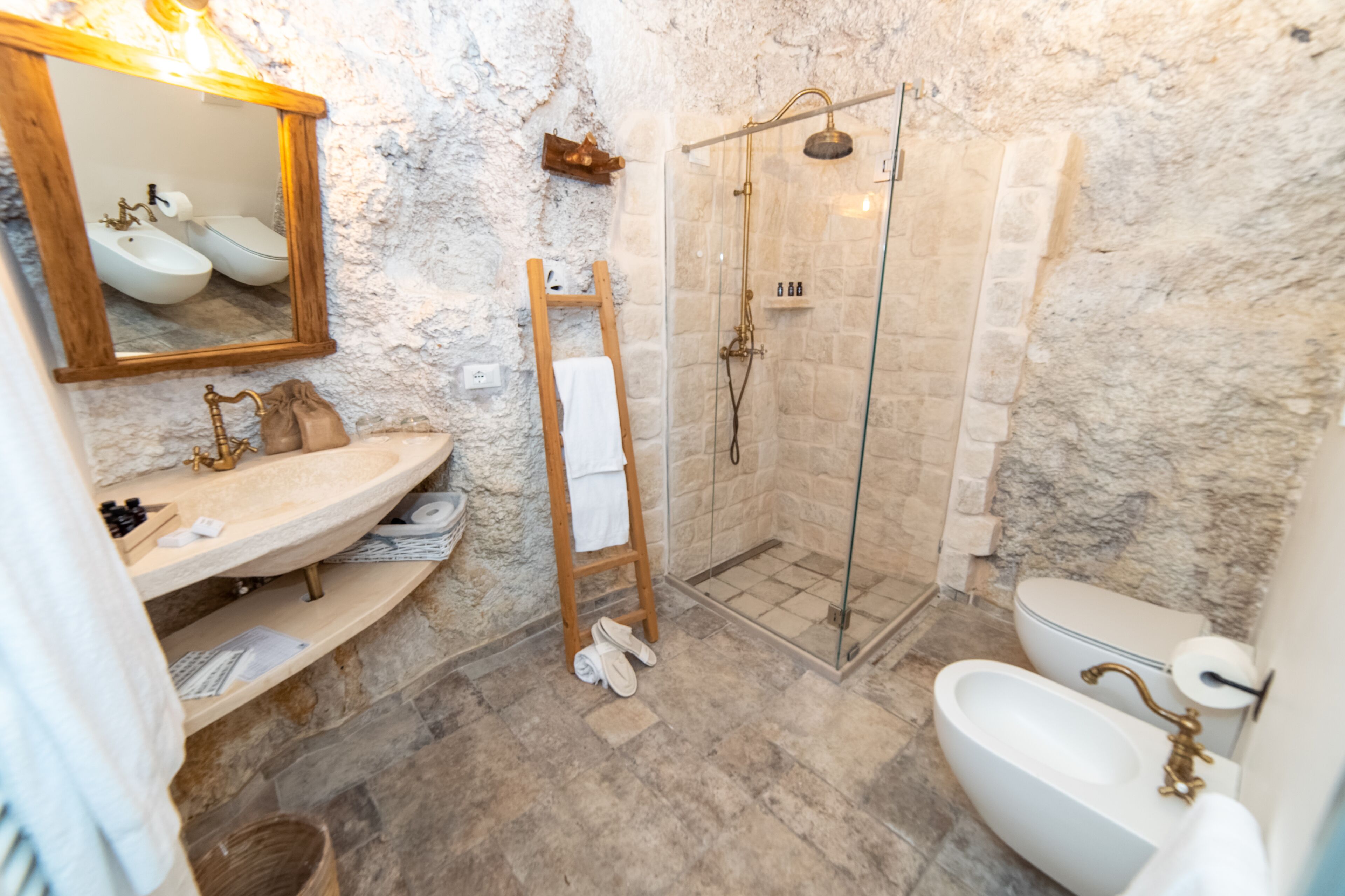 Un baño con pared de piedra, escalera de madera, cabina de ducha de vidrio y lavabo blanco.
