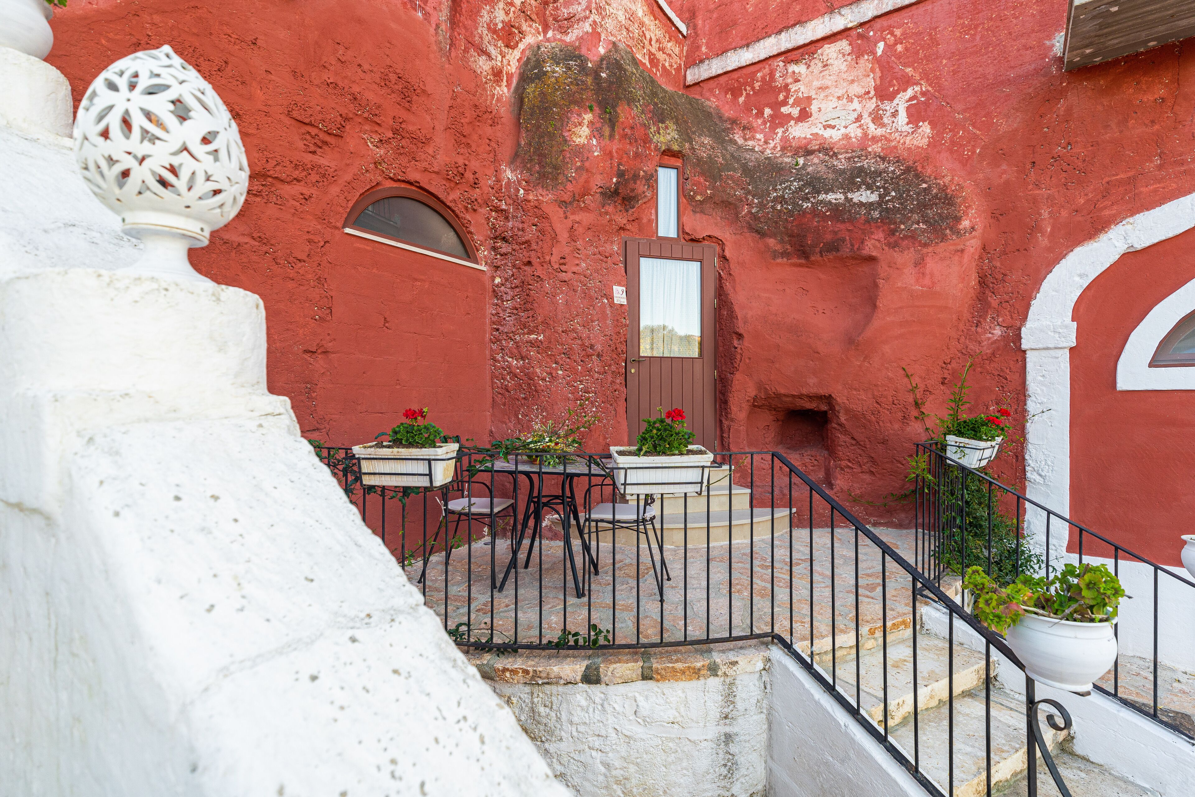 Un edificio rojo con detalles blancos, una pequeña escalera y mesas al aire libre con plantas en macetas.