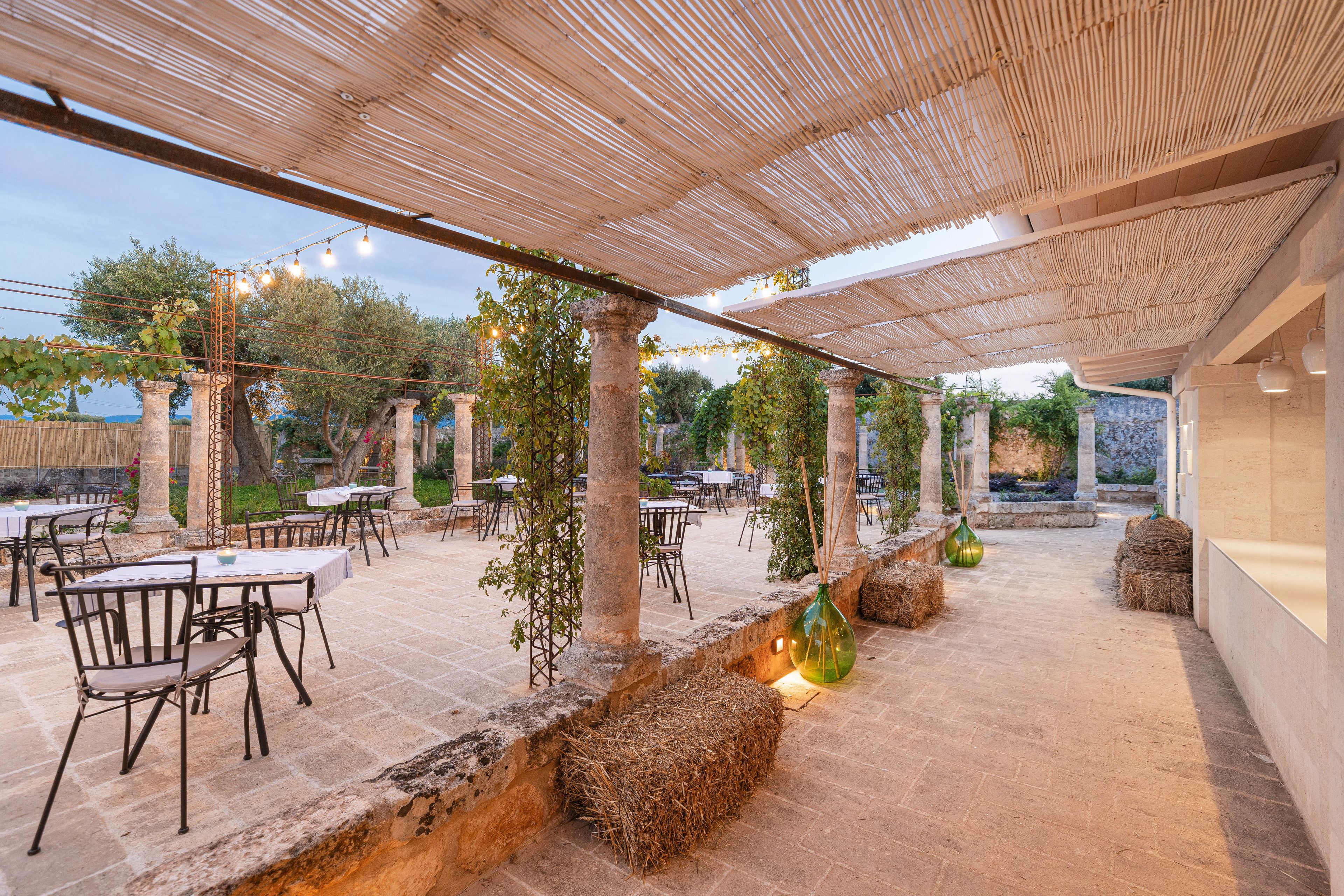 Un patio al aire libre con mesas y sillas, una pérgola con luces colgantes y un camino de piedra.