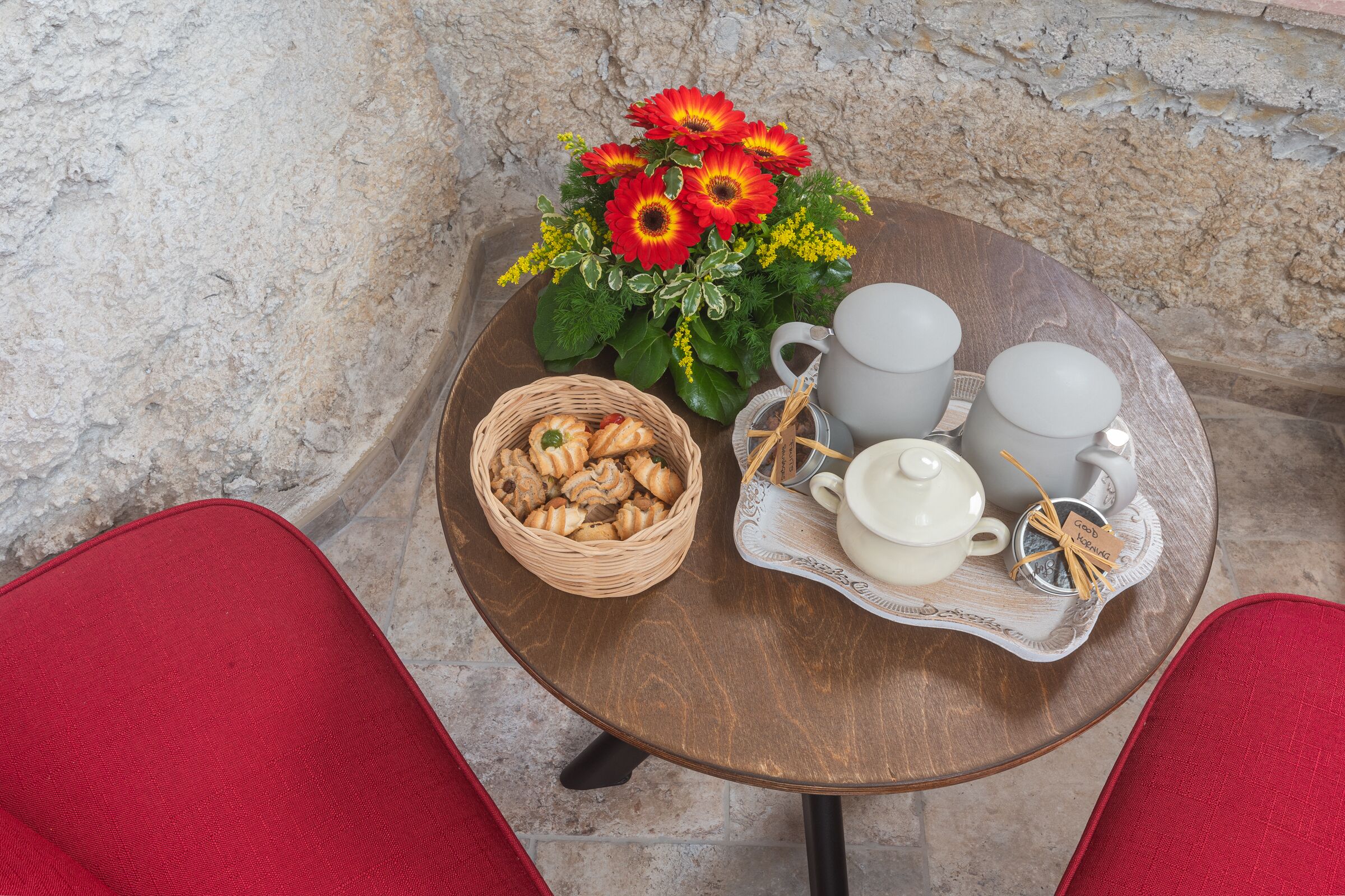 A round wooden table with a wicker basket of cookies, a tray with teacups and a teapot, and a bouquet of red and yellow flowers.