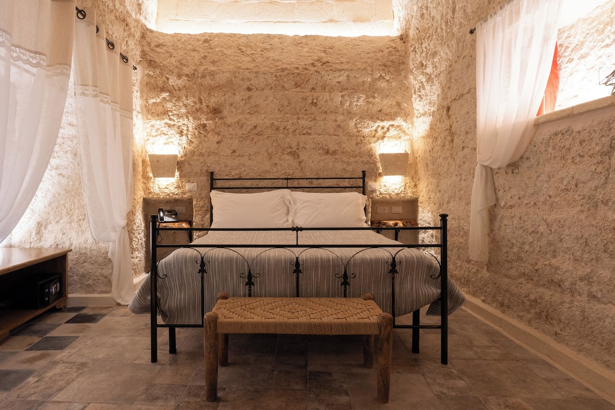 A bedroom with a stone wall, a four-poster bed, bedside lamps, a bench, and a wooden nightstand.