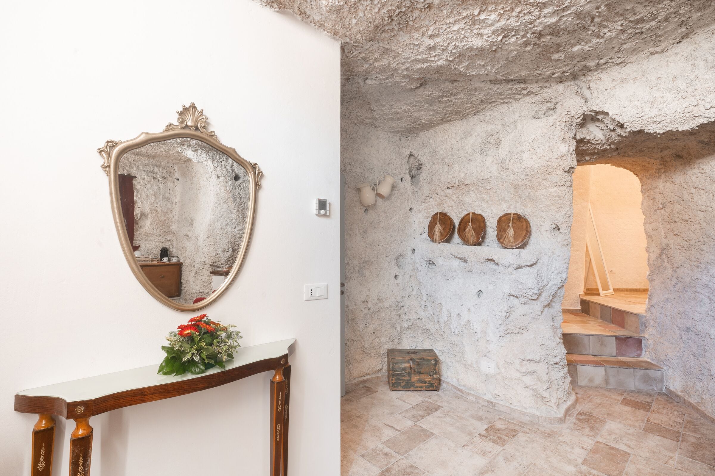 A room with a mirror, a wooden console table, a vase with flowers, and a stone wall with a wooden staircase.