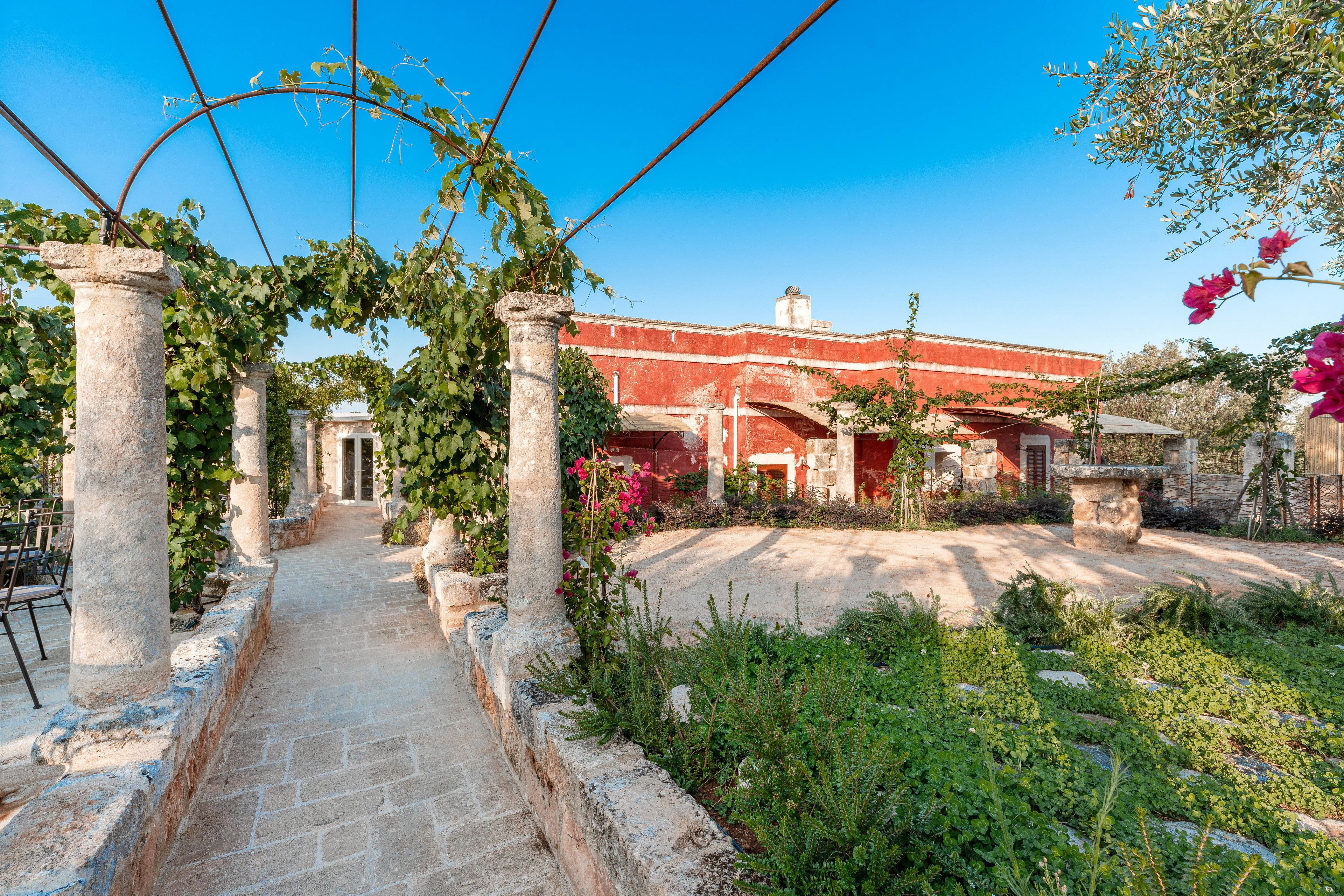 A pathway leading to a building with a red facade, surrounded by greenery and stone columns.