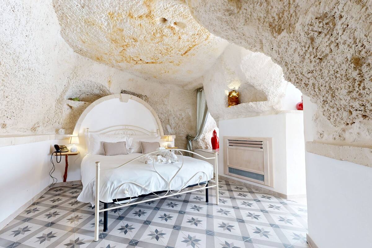 A bedroom with a white bed, patterned floor tiles, and a textured white wall.