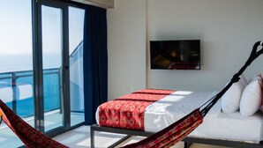 Et moderne hotelværelse med stor balkon, en seng med sengelagen i rødt og hvidt mønster, en hvid sofa og et fladskærms-tv monteret på væggen.