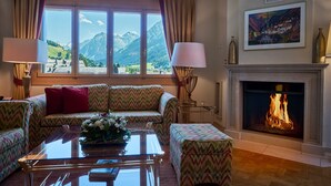 Ein gemütliches Wohnzimmer mit Sofa, Hocker und Couchtisch, einem Kamin und einem großen Fenster mit Blick auf die Berge.