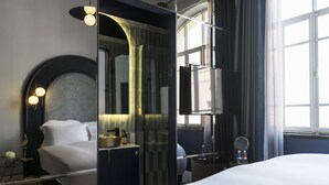 Habitación de hotel moderna con una cama grande, ducha envuelta en vidrio, tocador con espejo y vistas de la ciudad a través de amplias ventanas.