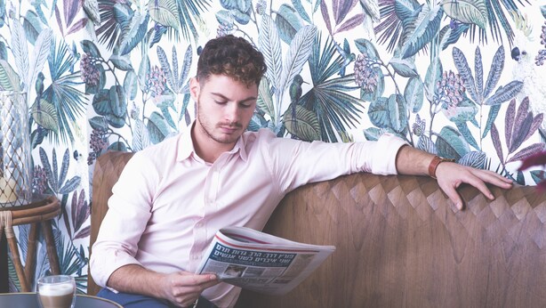 A person reading a newspaper on a cushioned bench with a floral wallpaper background.
