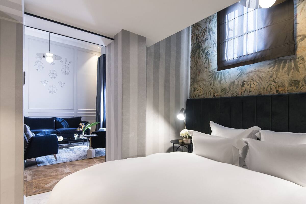 Une chambre d’hôtel moderne avec un grand lit, une tête de lit sombre et une vue sur un espace de vie comprenant un canapé et une table.