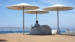 A person sitting on a large, curved bench under three large umbrellas on a wooden deck by the sea.