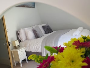 A neatly made bed with a white duvet, grey pillows, and a rolled white towel. There is a small bedside table with a box and a lamp. The room has a light-colored carpet and a partial view of a door frame. - Broadpark Villa (Ilfracombe)