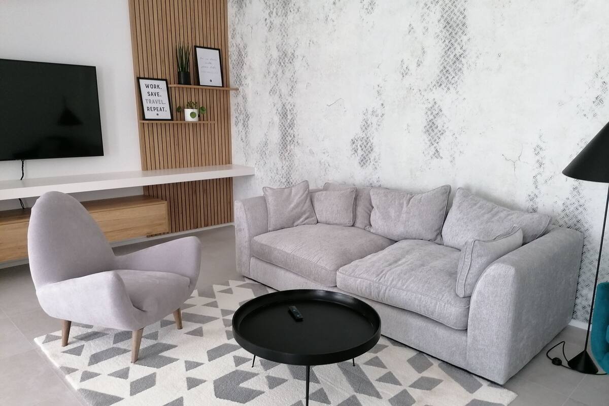 Una sala de estar moderna con un sofá gris, una mesa de centro negra redonda y un sillón gris claro. Un televisor de pantalla plana está montado en la pared, y hay dos citas enmarcadas en un estante de madera.