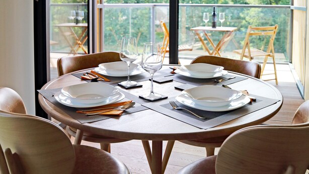 A dining area with a round table set for four, surrounded by wooden chairs. A large window provides a view of the outdoors.
