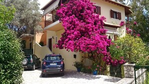 Una casa de dos pisos con balcón, coches estacionados y un vibrante arbusto de buganvilla.