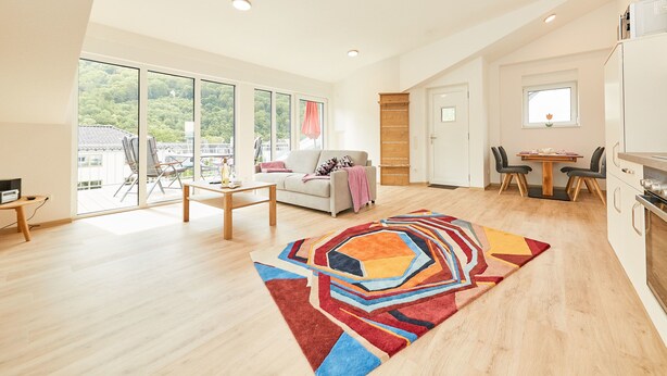 Een ruime woonkamer met een groot raam, een bank, een bijzettafeltje en een kleurrijk vloerkleed.