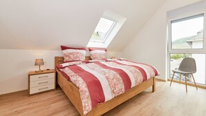 Een slaapkamer met een schuine kap, een bed met een bloemenprintdeken, een nachtkastje met een lamp en een stoel bij een groot raam.